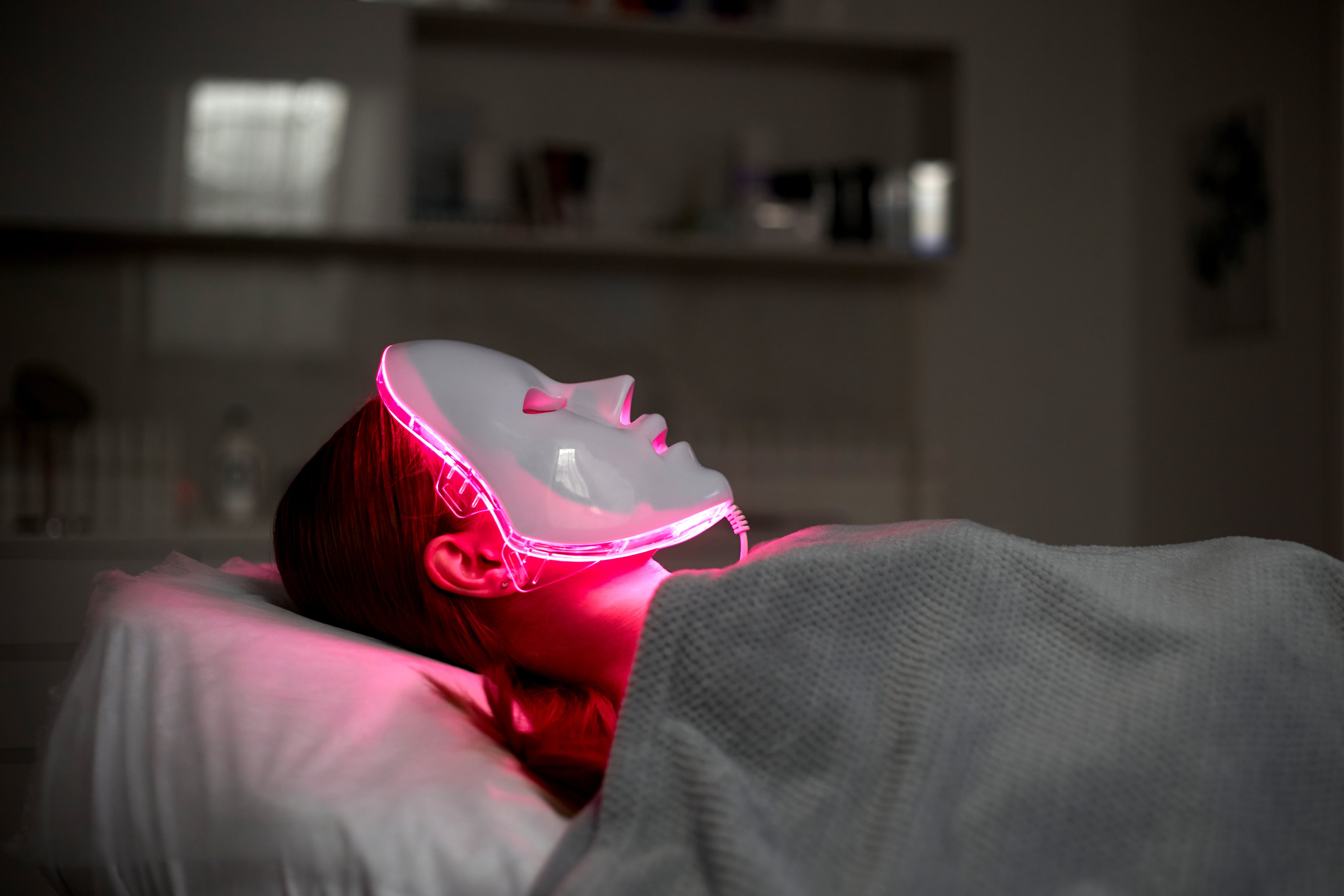 A woman is lying on a spa bed under a towel with an led mask on her face
