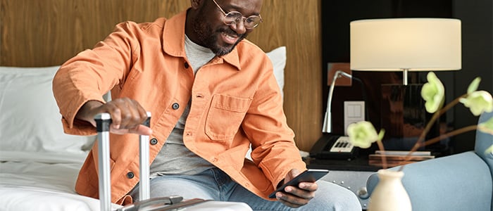 Hotel guest sitting on a bed in a hotel room smiling and looking at mobile phone