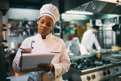 Female chef standing in kitchen, taking notes with a notepad.