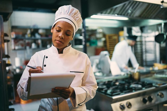 Female chef standing in kitchen, taking notes with a notepad.